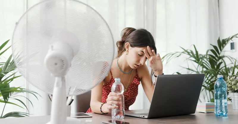 A hot exhausted woman sits in front of a fan drinking water