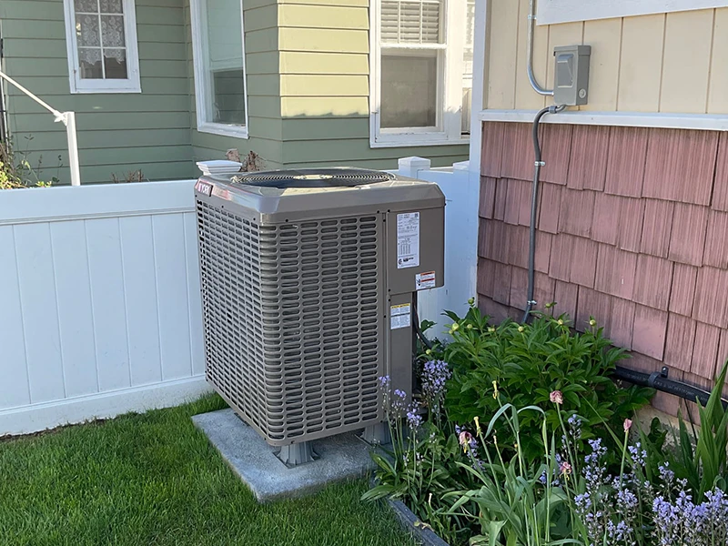 An air conditioning condenser professionally installed. It is in a green yard by a white fence.